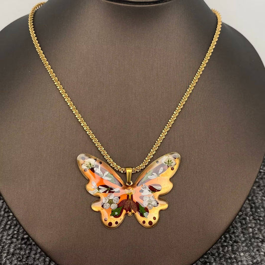 The Mio Queena - Dried Flower Brass Butterfly 18K Golden SS Necklace features a butterfly-shaped resin pendant in orange, brown, and gold hues, adorned with delicate pressed flowers and suspended from a gold-plated chain. This elegant necklace by Mio Queena is beautifully displayed on a brown jewelry stand.