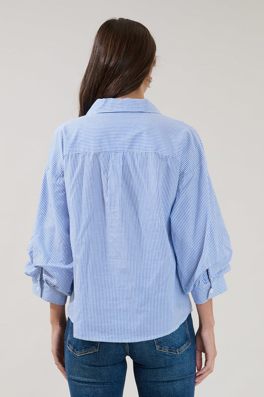 A person with long brown hair, seen from behind, wears the Sugarlips Chalet Button Down Stripe Top by Sugarlips—with blue and white stripes and rolled sleeves—paired with blue jeans against a plain background.