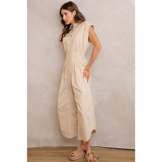 A woman stands in profile against a plain light-colored wall and wooden floor, wearing the So Me SLEEVELESS WIDE LEG STRIPE JUMPSUIT by So Me. The beige and white stripe jumpsuit features an elastic waist, wide legs, and is paired with beige sandals.