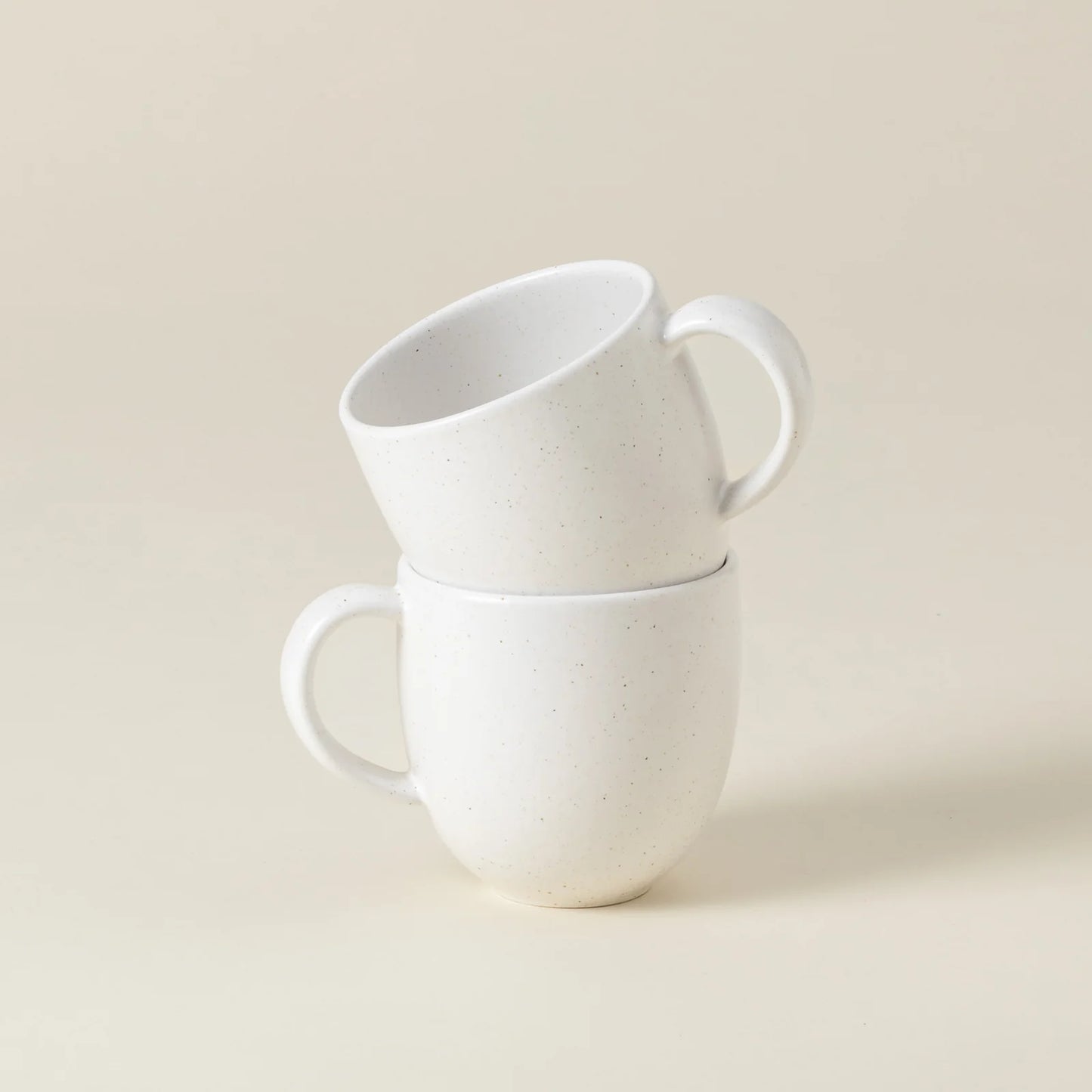 Two Casafina “Pacifica - White” mugs by Casafina are stacked, with one mug resting inside and slightly tilted on top of the other, set against a plain beige background.