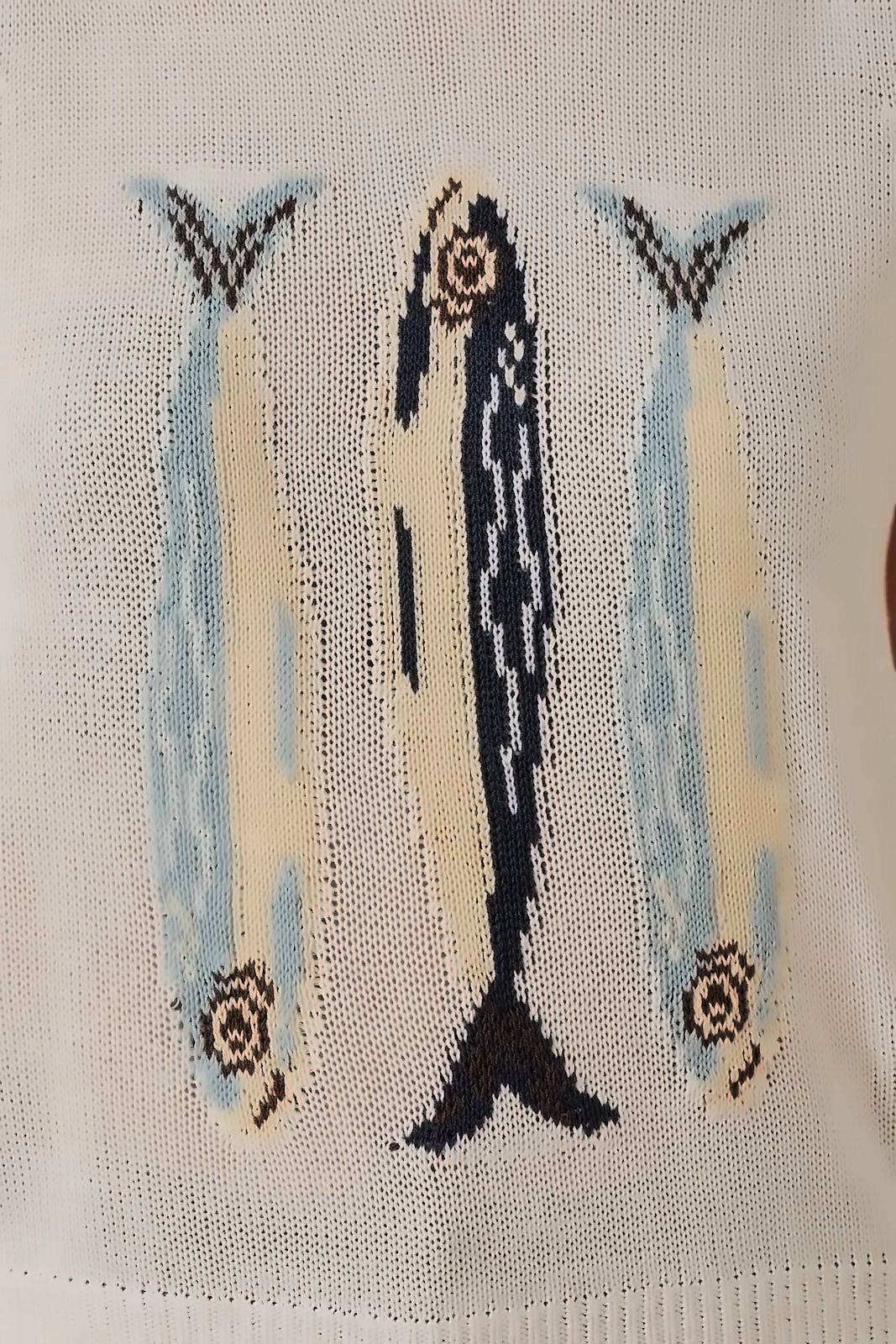 A close-up of a size small Sunday Edition by LE LIS ROUND NECK 'FISH' GRAPHIC SWEATER VEST, featuring knitted fabric with three stylized fish in blue, beige, and black, oriented vertically with their heads at the bottom and tails at the top. Model is 5'10".