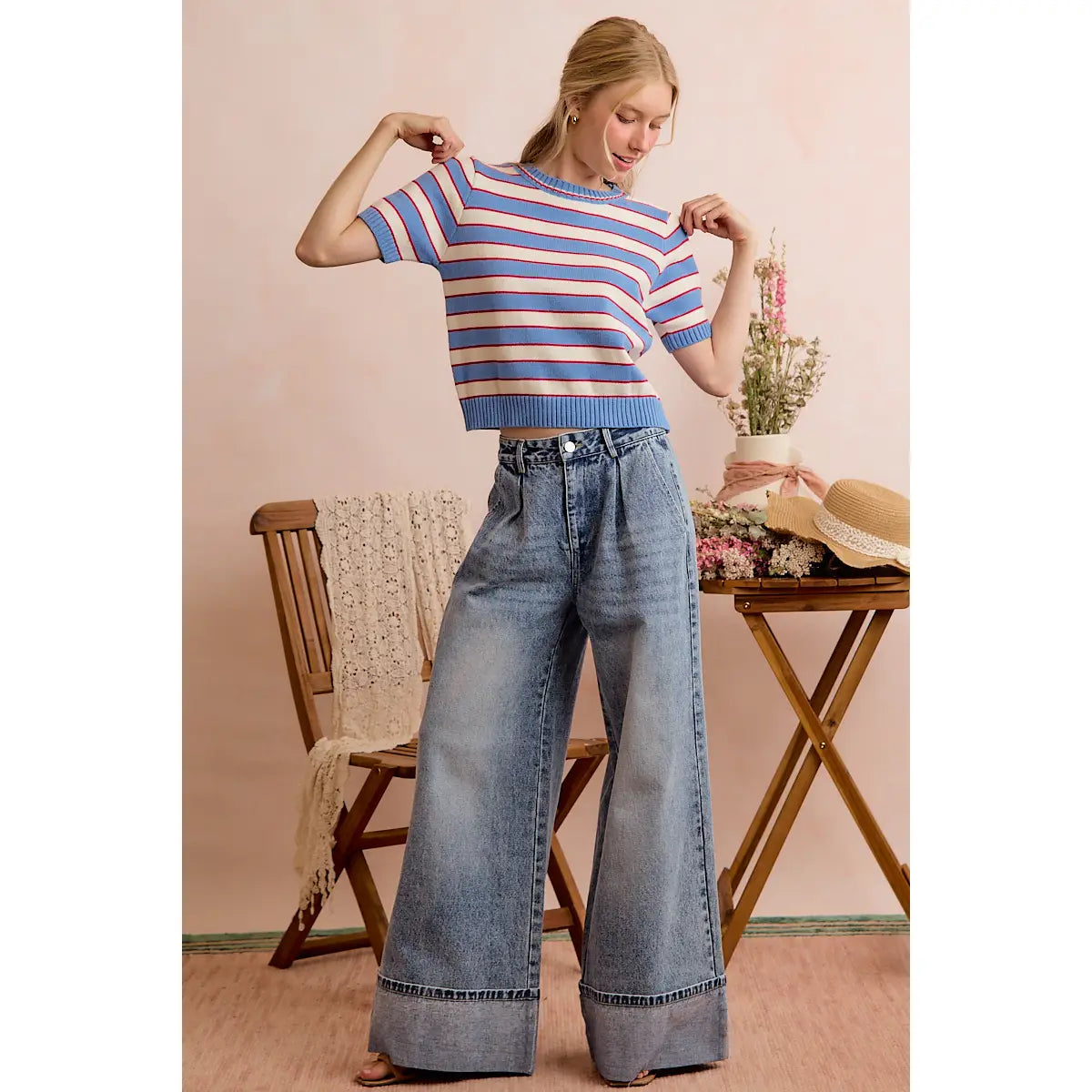 A woman poses indoors wearing the So Me Stripe Knit Crew Neck Short Sleeve Sweater Top by So Me, paired with high-waisted wide-leg jeans. She stands near a wooden chair draped with a lace throw, beside a table adorned with flowers and a straw hat.