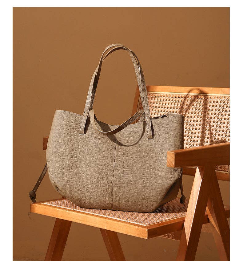 A Parker & Hyde Brooklyn Tote in Oyster leather with double handles rests elegantly on a light wooden chair featuring a woven cane backrest, set against a warm brown background.