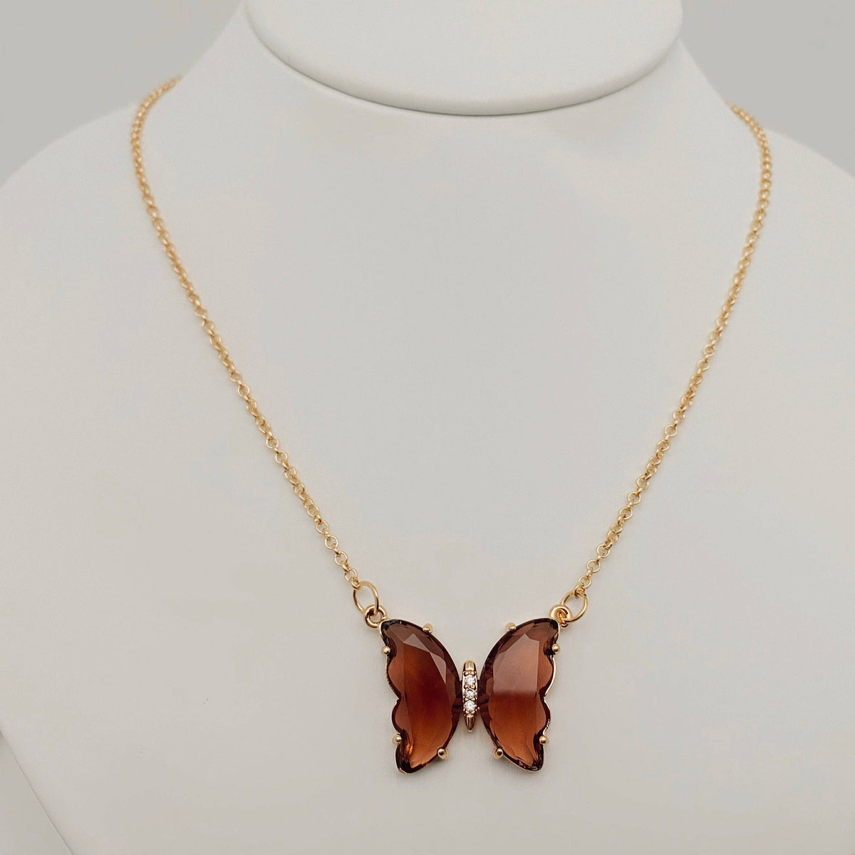The Mio Queena - Multicolor Glass Butterfly Pendant Necklace by Mio Queena features a butterfly-shaped glass pendant in translucent brown, suspended from a gold chain and elegantly displayed on a white jewelry stand.