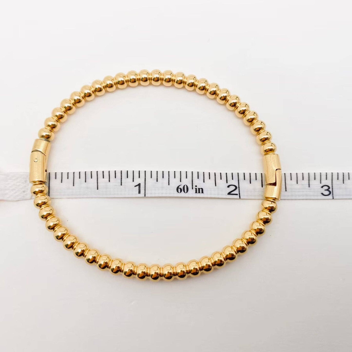 A Mio Queena - 4mm Beads Gold Plated Stainless Steel Snap Bangle (FGS) rests on a white surface, with a measuring tape indicating its approximately 2.5-inch diameter—a stunning jewelry gift from Mio Queena.