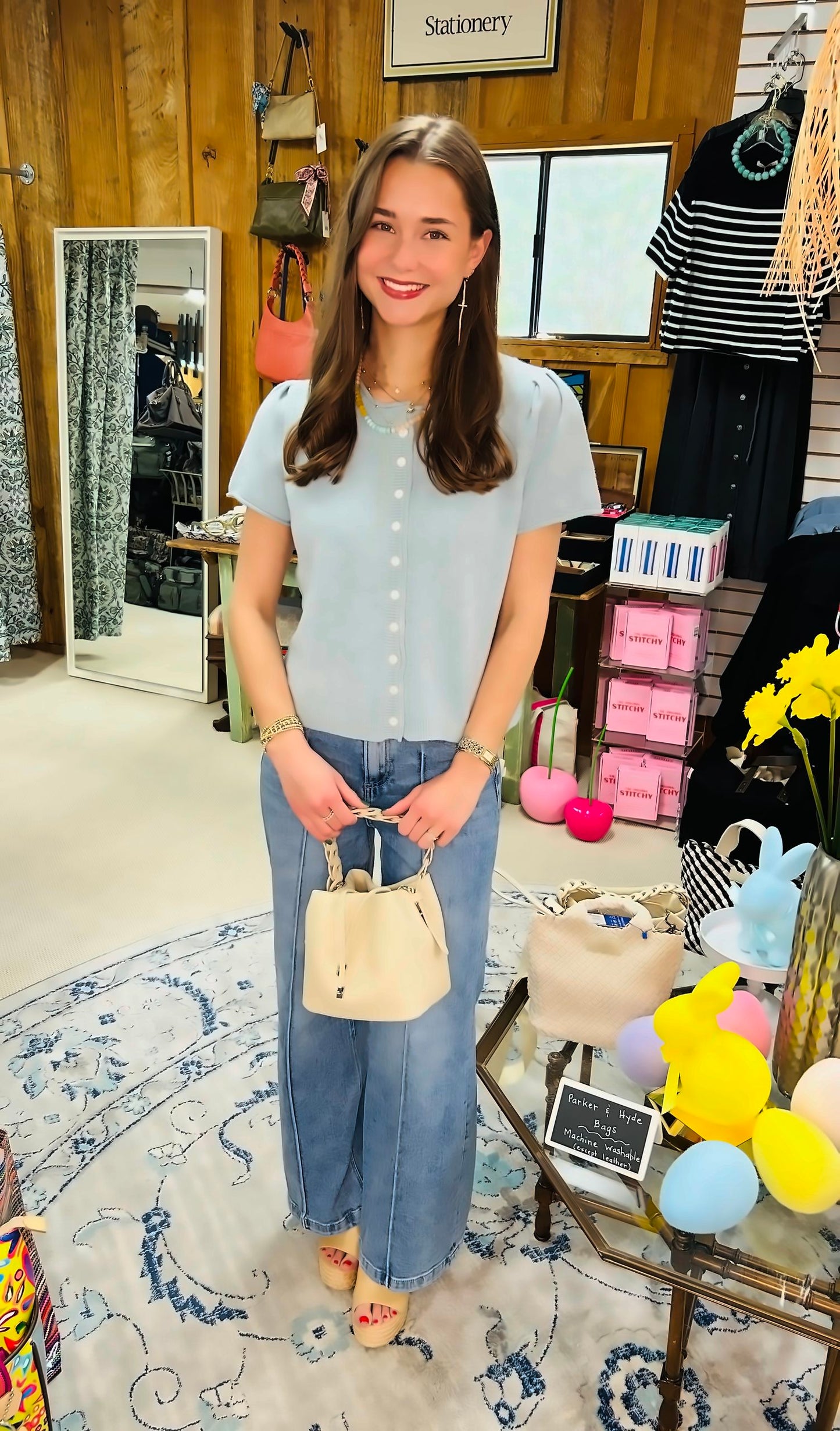 A smiling woman wearing the &MERCI Short Sleeve Sweater Cardigan stands in a boutique, holding a small cream handbag. She is surrounded by colorful decor, baskets, and flowers.