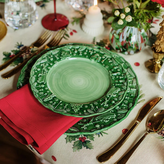 A festive table setting featuring the Vietri Fiabe Di Natale Green Dinner Plate, complemented by a handpainted holiday plate, a red napkin, gold cutlery, and a holiday-themed tablecloth. A small candle and floral arrangement complete the holiday decor.
