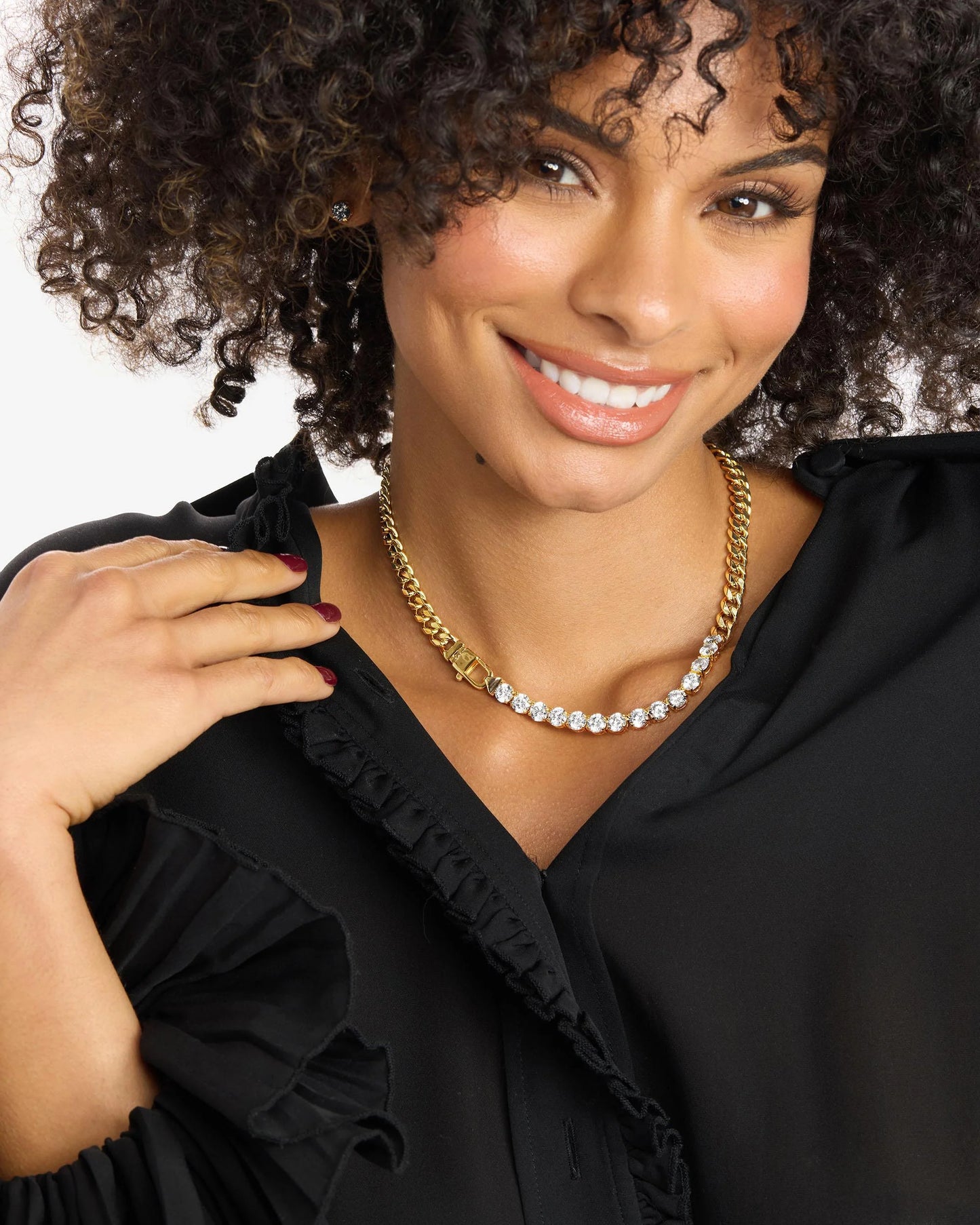 Smiling woman with curly hair wears a black ruffled blouse and two necklaces—one a striking 18k gold plated Cuban chain, the other the Melinda Maria Julian Really Loves Diamonds Necklace 16" adorned with clear simulated diamonds—posing with one hand near her collar.