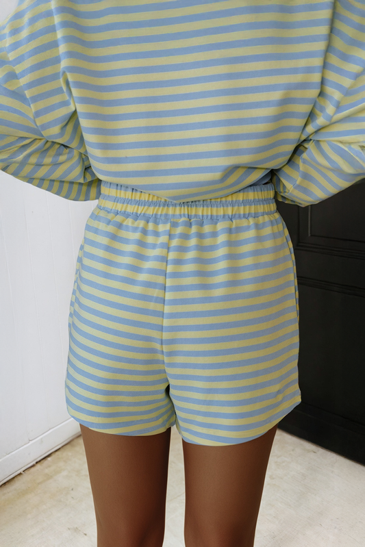 A person is seen standing with hands on hips, facing away from the camera, wearing the LA Society - LAS Stripe Color Block Pullover Shorts 2pcs Set—a matching light blue and yellow horizontally striped loungewear outfit crafted in soft cotton from LA Society.