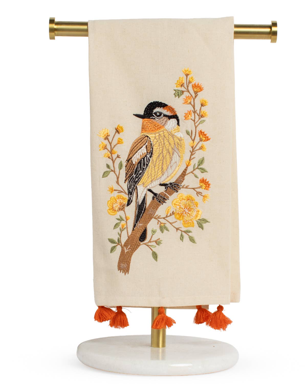 The Royal Standard - Autumn Sparrow Hand Towel features a cream-colored fabric adorned with an embroidered sparrow resting on a branch accented by yellow flowers, while orange tassels embellish the bottom edge. Displayed over a gold and marble stand, this hand towel from The Royal Standard brings a charming touch to your kitchen décor.