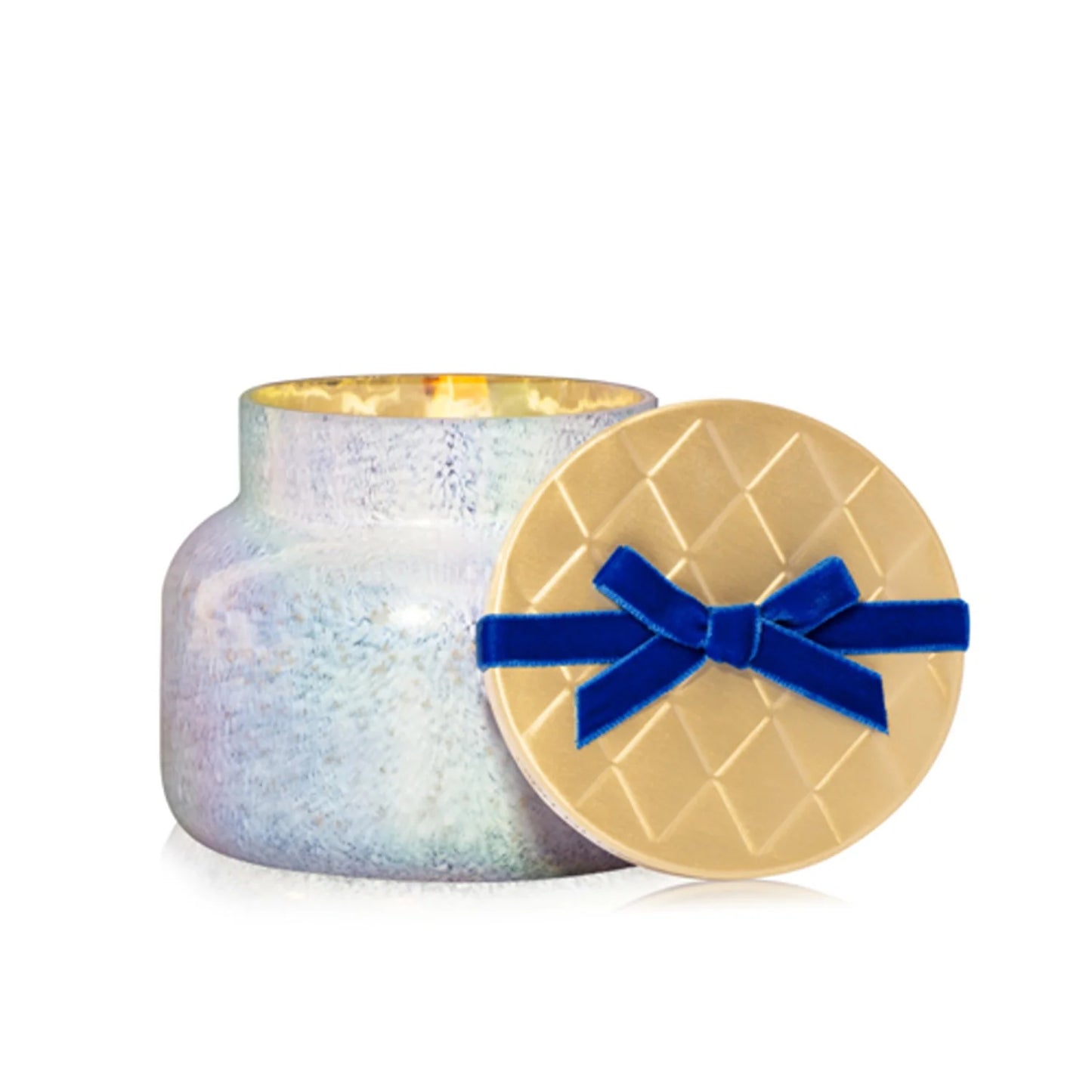 A glowing Capri Blue Volcano Winter Bazaar Signature Candle sits inside a shimmering iridescent glass jar, with the gold, quilt-patterned lid and a blue ribbon bow resting beside it.
