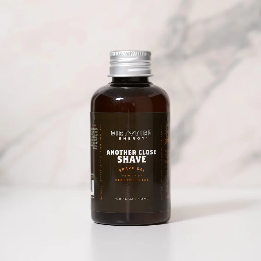 A 4.8 fl oz brown bottle of Dirty Bird Another Close Shave Gel, featuring bentonite clay shave gel and labeled with the Dirty Bird brand, sits on a white surface against a light gray marbled background.
