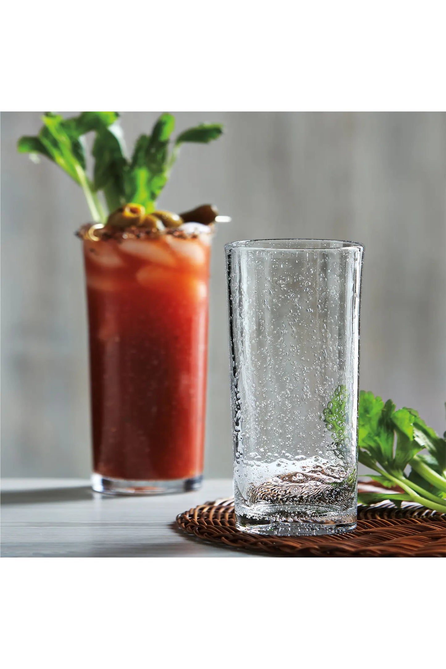 A Tag "Bubble" Tall Bloody Mary Glass by Tag sits empty on a table next to a garnished Bloody Mary cocktail and some fresh celery, with a woven mat partially visible underneath the glass.