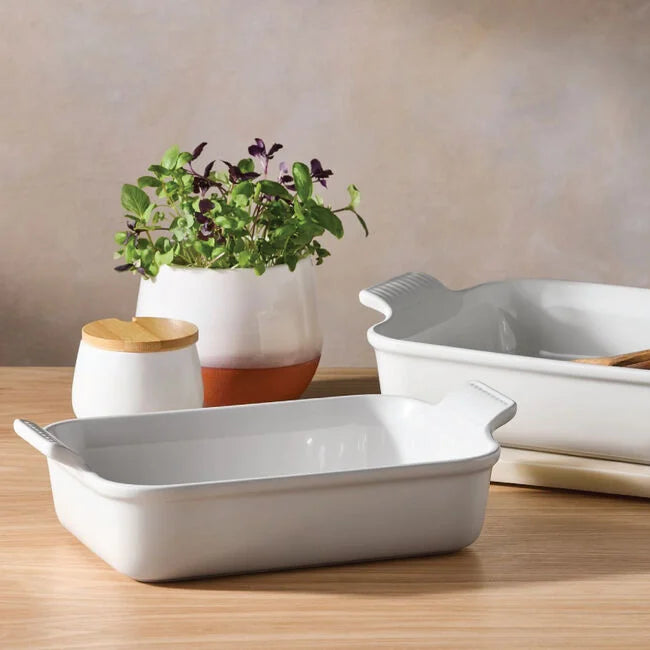 Two Le Creuset 2.5 qt Heritage Rectangular Baking Dishes in white rest on a wooden table, with a small potted plant and a white jar with a wooden lid in the background.