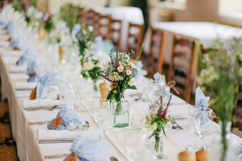 A long banquet table is set with floral centerpieces, wine glasses, cutlery, and blue checkered napkins. Wooden chairs line both sides, creating an elegant setting for a formal meal or celebration.