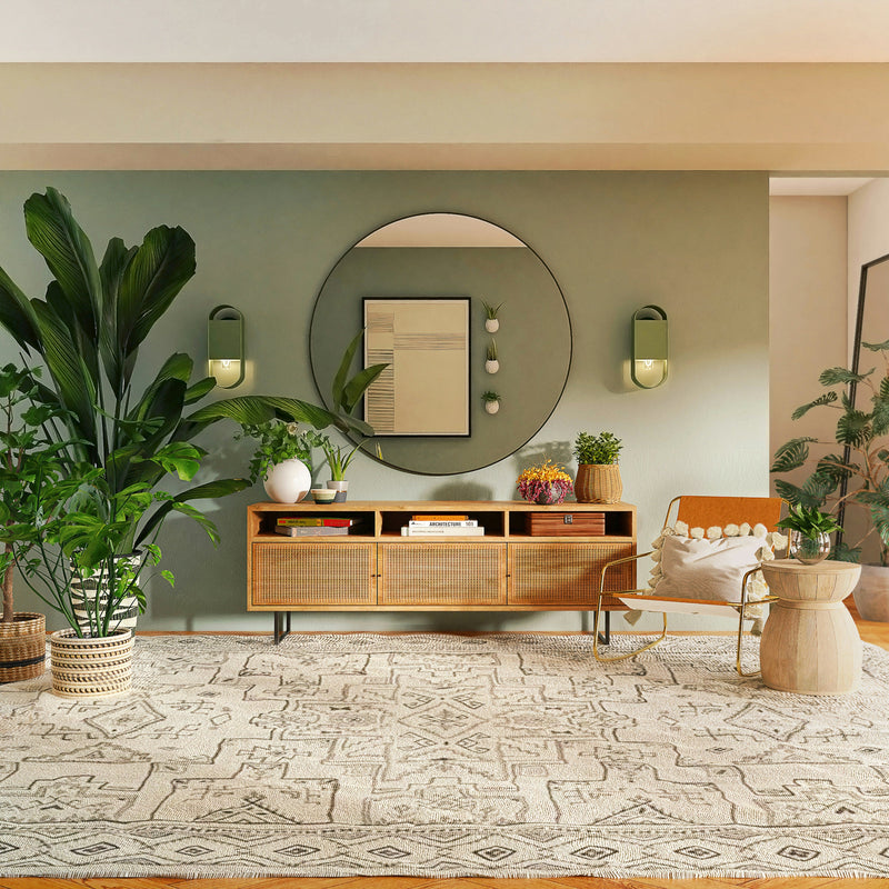Bright living room with a large plant, woven sideboard, chair, round mirror, wall sconces, and decorative items on a patterned rug over light wood flooring.