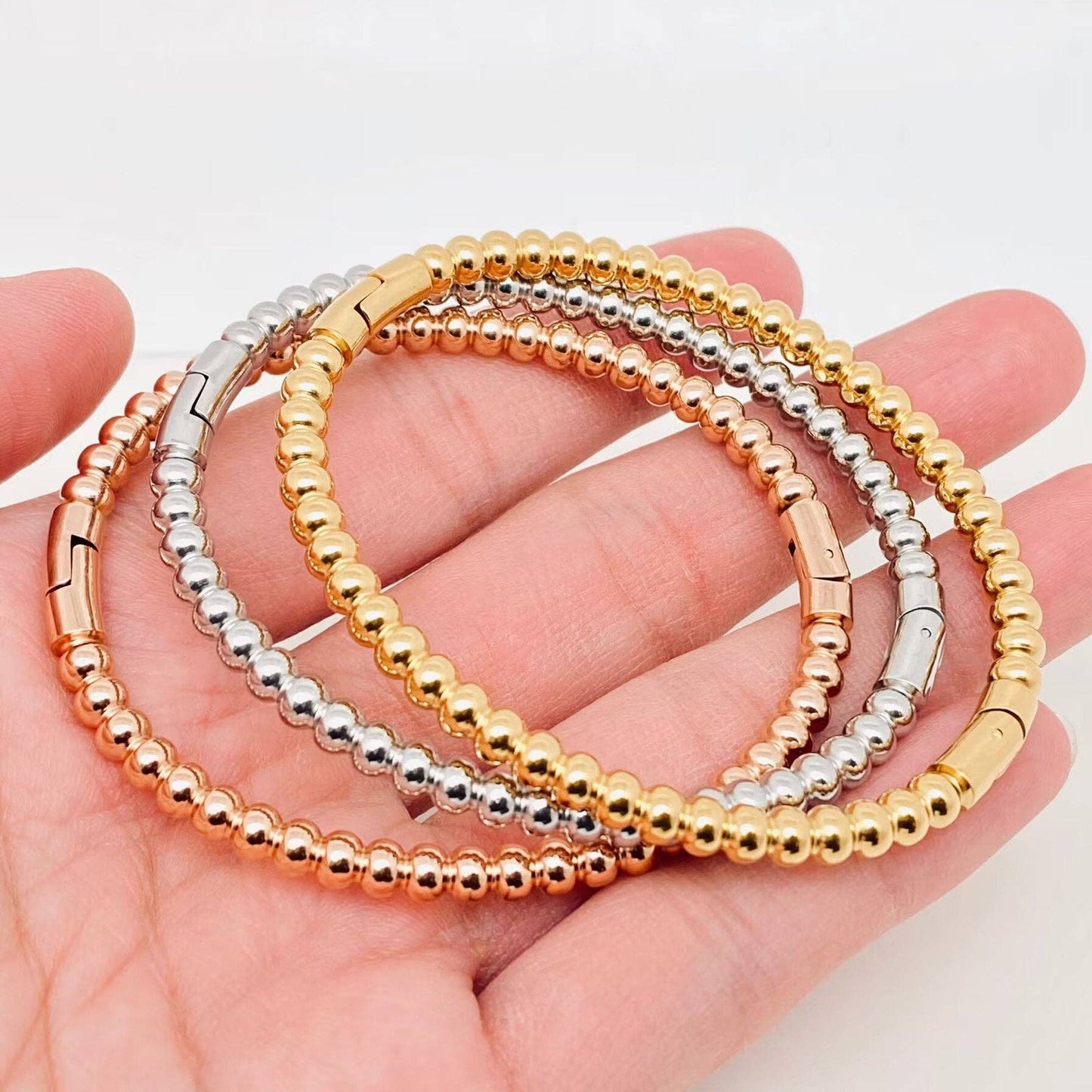 A hand holds three Mio Queena - 4mm Beads Gold Plated Stainless Steel Snap Bangles, each crafted from durable stainless steel and finished in elegant gold, silver, and rose gold tones, displayed against a white background. These 18K gold plated pieces by Mio Queena make perfect jewelry gifts.