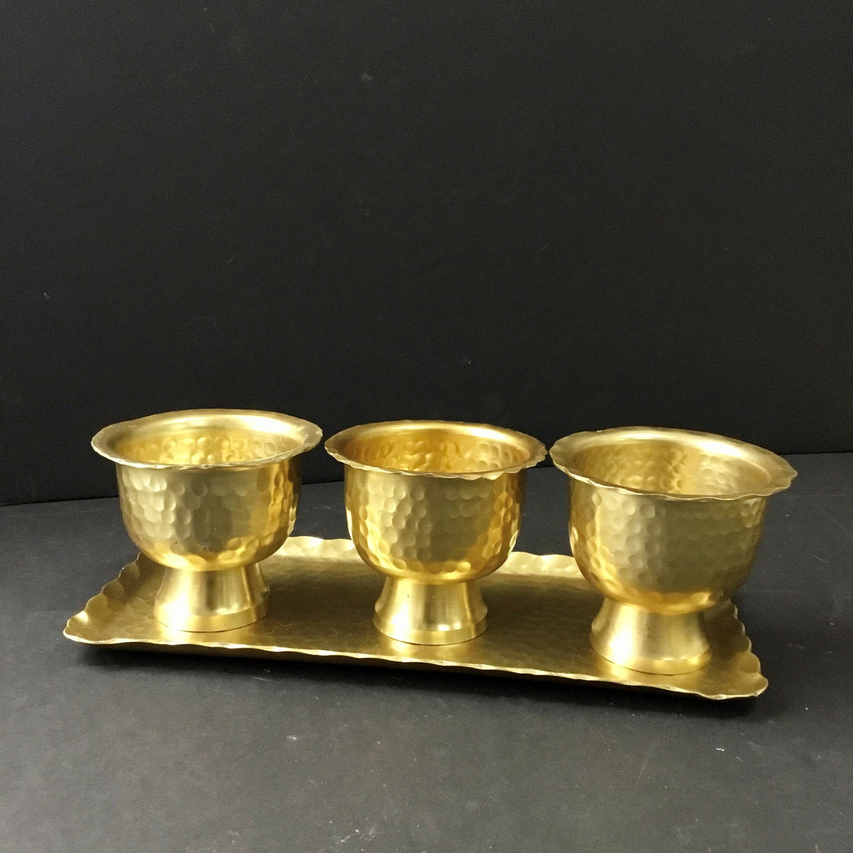 Three gold hammered aluminum pedestal bowls on matching rectangular tray, side view against dark background