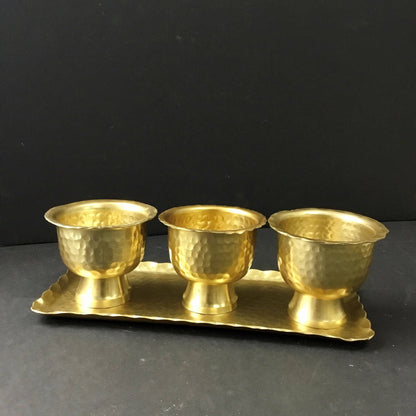 Three gold hammered aluminum pedestal bowls on matching rectangular tray, side view against dark background