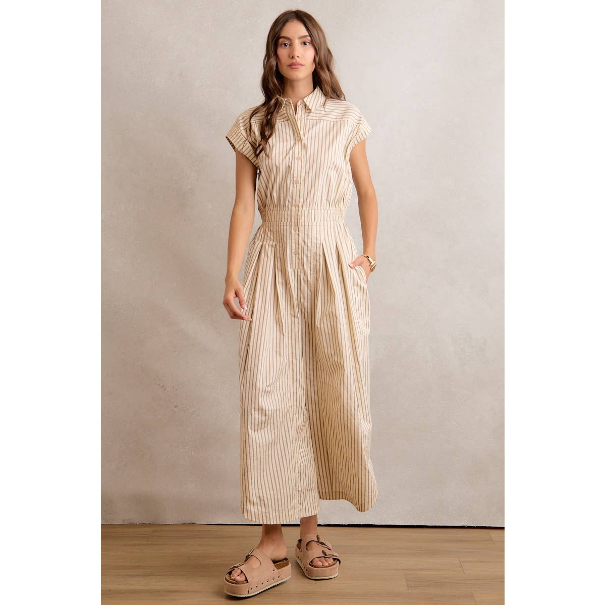 A woman with long hair and a neutral expression stands against a light beige wall, wearing the So Me Sleeveless Wide Leg Stripe Jumpsuit in tan and white stripes, paired with tan platform sandals.