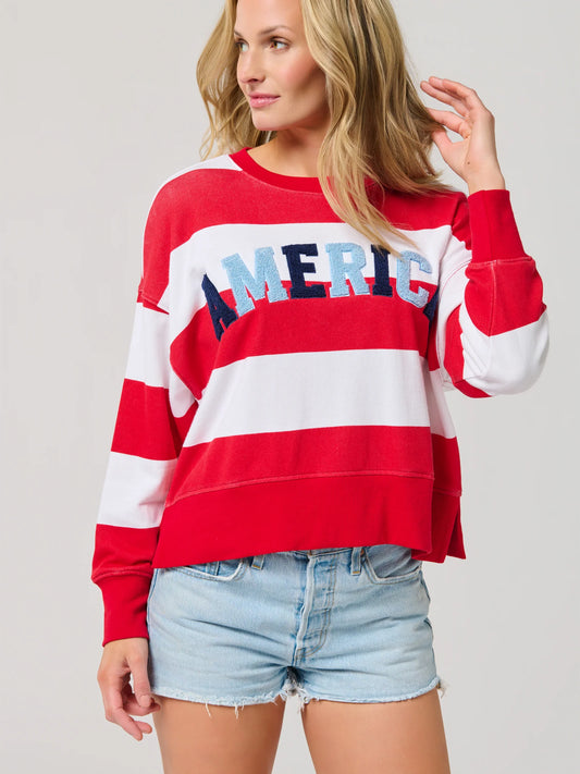 A woman stands against a plain background, touching her hair and smiling slightly while wearing the Shiraleah "America" Rugby Stripe Relaxed Sweatshirt in white and light denim shorts.