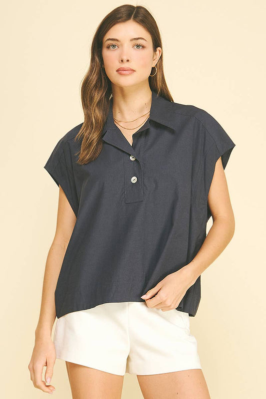 A woman with long brown hair stands against a beige background wearing the PINCH - SOLID CAP SLEEVE BLOUSE TOP by PINCH, a navy button-up blouse featuring classic collar and cap sleeves, paired with white shorts. She accessorizes minimally and has a neutral expression, with one hand resting on her hip.