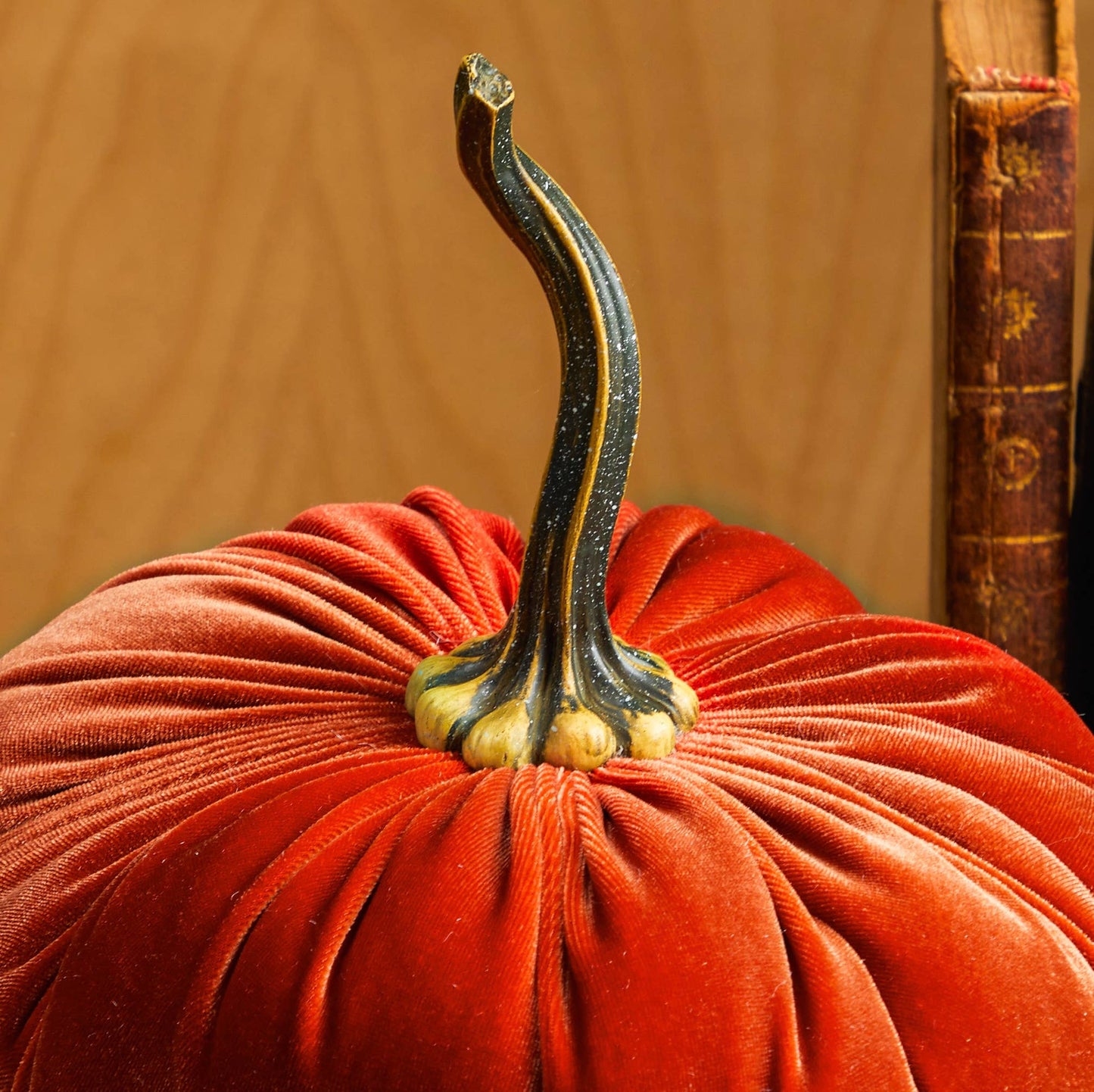 A close-up of Your Heart’s Content - Pumpkin Extra Large Velvet, crafted from plush orange velvet with a lifelike faux stem, is displayed beside a vintage book against a wood background—making it an elegant centerpiece for weddings or charming holiday mantel décor.