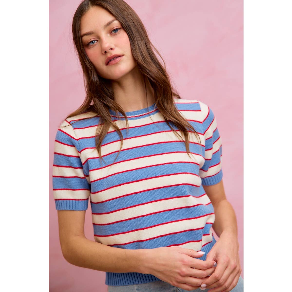 A woman with long brown hair models the So Me STRIPE KNIT CREW NECK SHORT SLEEVES SWEATER TOP, standing against a pale pink background. This trendy sweater by So Me features a crew neck, short sleeves, and bold blue, white, and red stripes for a fresh, stylish look.
