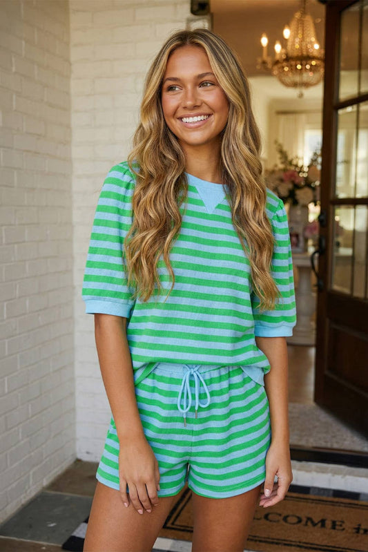 A smiling woman with long, wavy hair wears the LA Society LAS Stripe Puff Sleeve T Shirt Matching Shorts Set in green and light blue stripes, standing in front of an open door with a chandelier and flowers visible in the background.