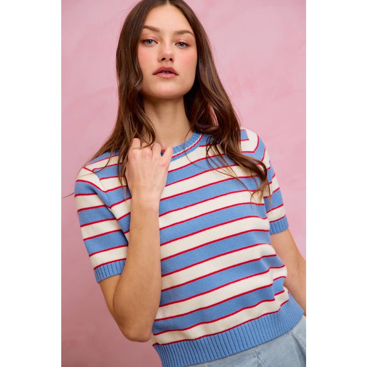 A woman with long brown hair stands confidently against a pink background, wearing the So Me STRIPE KNIT CREW NECK SHORT SLEEVES SWEATER TOP—a trendy cream-colored sweater featuring bold red and blue stripes—from the brand So Me.