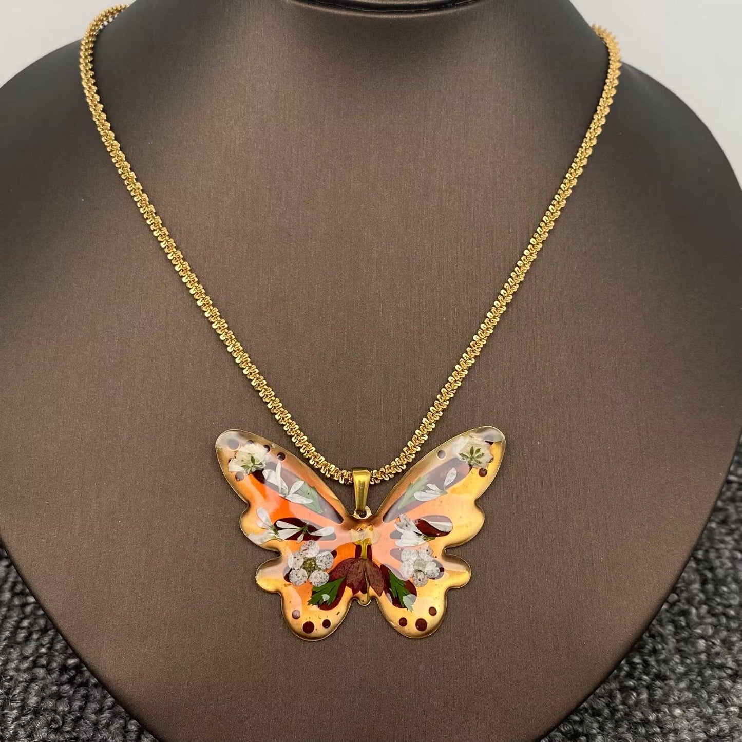 The Mio Queena - Dried Flower Brass Butterfly 18K Golden SS Necklace features a butterfly-shaped resin pendant in orange, brown, and gold hues, adorned with delicate pressed flowers and suspended from a gold-plated chain. This elegant necklace by Mio Queena is beautifully displayed on a brown jewelry stand.