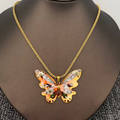 The Mio Queena - Dried Flower Brass Butterfly 18K Golden SS Necklace features a butterfly-shaped resin pendant in orange, brown, and gold hues, adorned with delicate pressed flowers and suspended from a gold-plated chain. This elegant necklace by Mio Queena is beautifully displayed on a brown jewelry stand.
