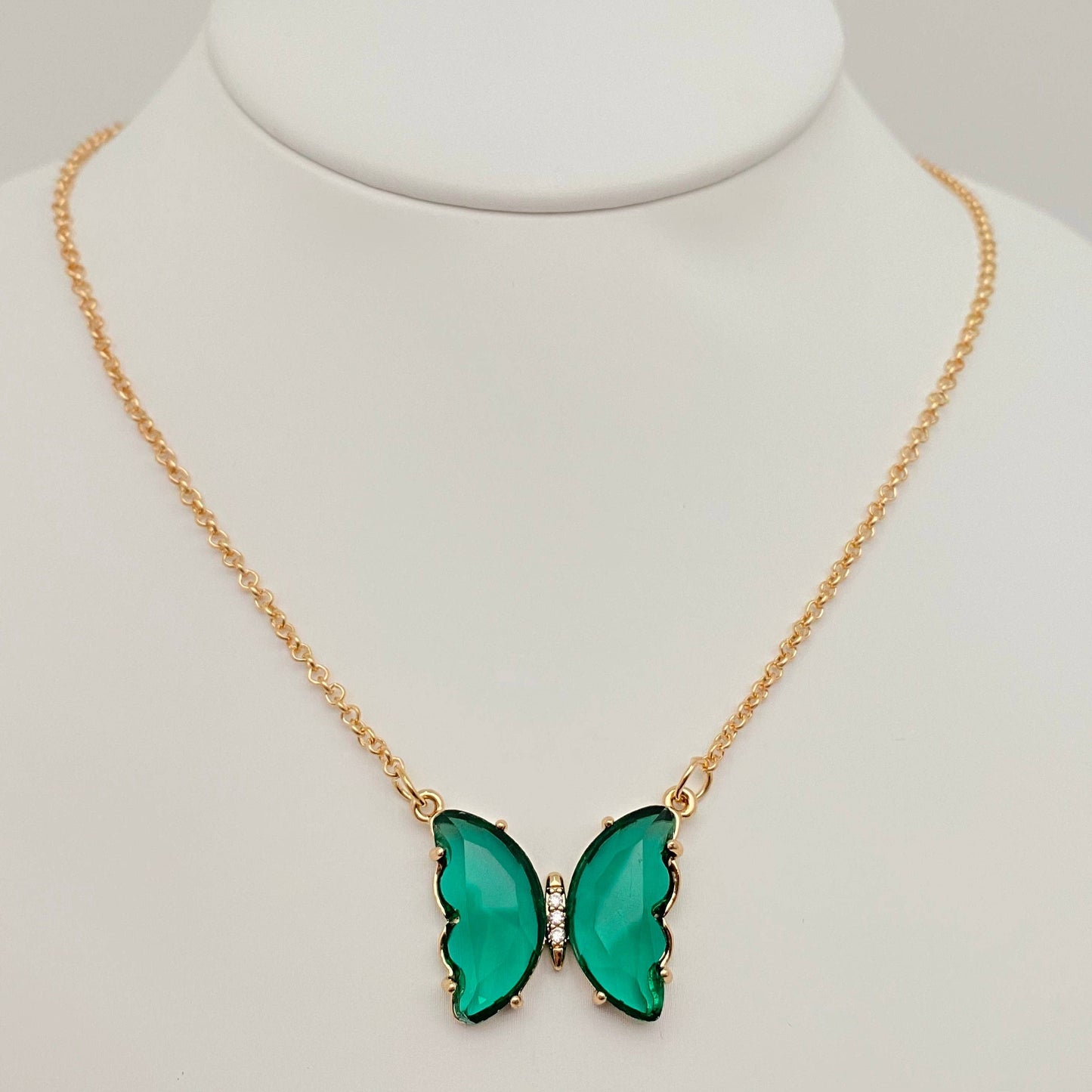 Introducing the Mio Queena - Multicolor Glass Butterfly Pendant Necklace by Mio Queena, featuring a stunning green butterfly pendant accented with small clear stones at its center. Displayed on a white jewelry stand, this hypoallergenic necklace combines elegance and comfort, making it suitable for sensitive skin.