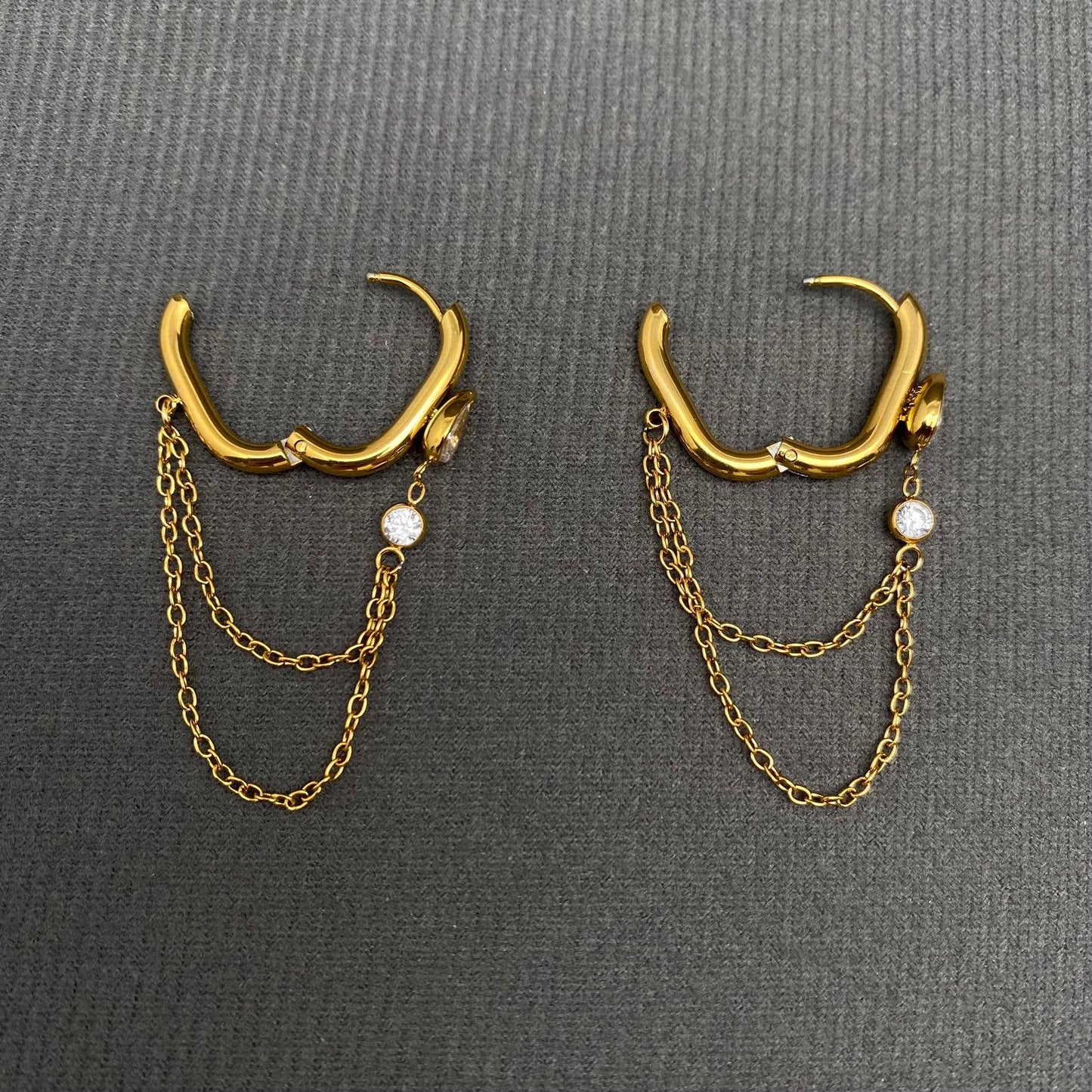 Mio Queena's Chain Fringe Zicon Inlaid 18K Gold Plated Stainless Steel Earrings (FGS) feature gold hoops with double chain dangles and sparkling small round gemstones, elegantly displayed on a textured dark gray surface. Expertly crafted from non-tarnish, 18K gold plated stainless steel, these Mio Queena earrings offer both timeless style and exceptional durability.
