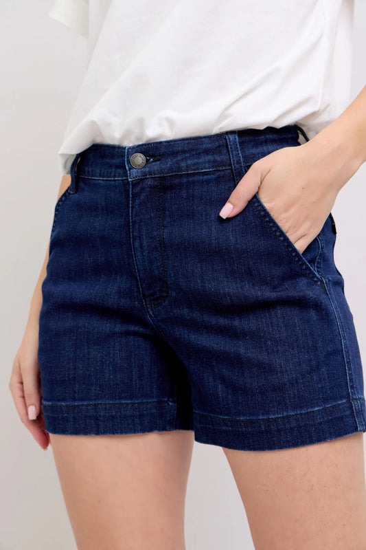 A person is wearing Judy Blue Dark Wash Denim High Waist Shorts by Judy Blue, paired with a white t-shirt and one hand in their pocket. The photo is cropped from mid-torso to mid-thigh.
