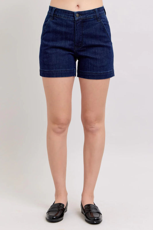 A person wearing Judy Blue Dark Wash Denim High Waist Shorts by Judy Blue and black loafers stands against a plain light gray background. The image is cropped at the waist and mid-calf, highlighting the shorts and shoes.