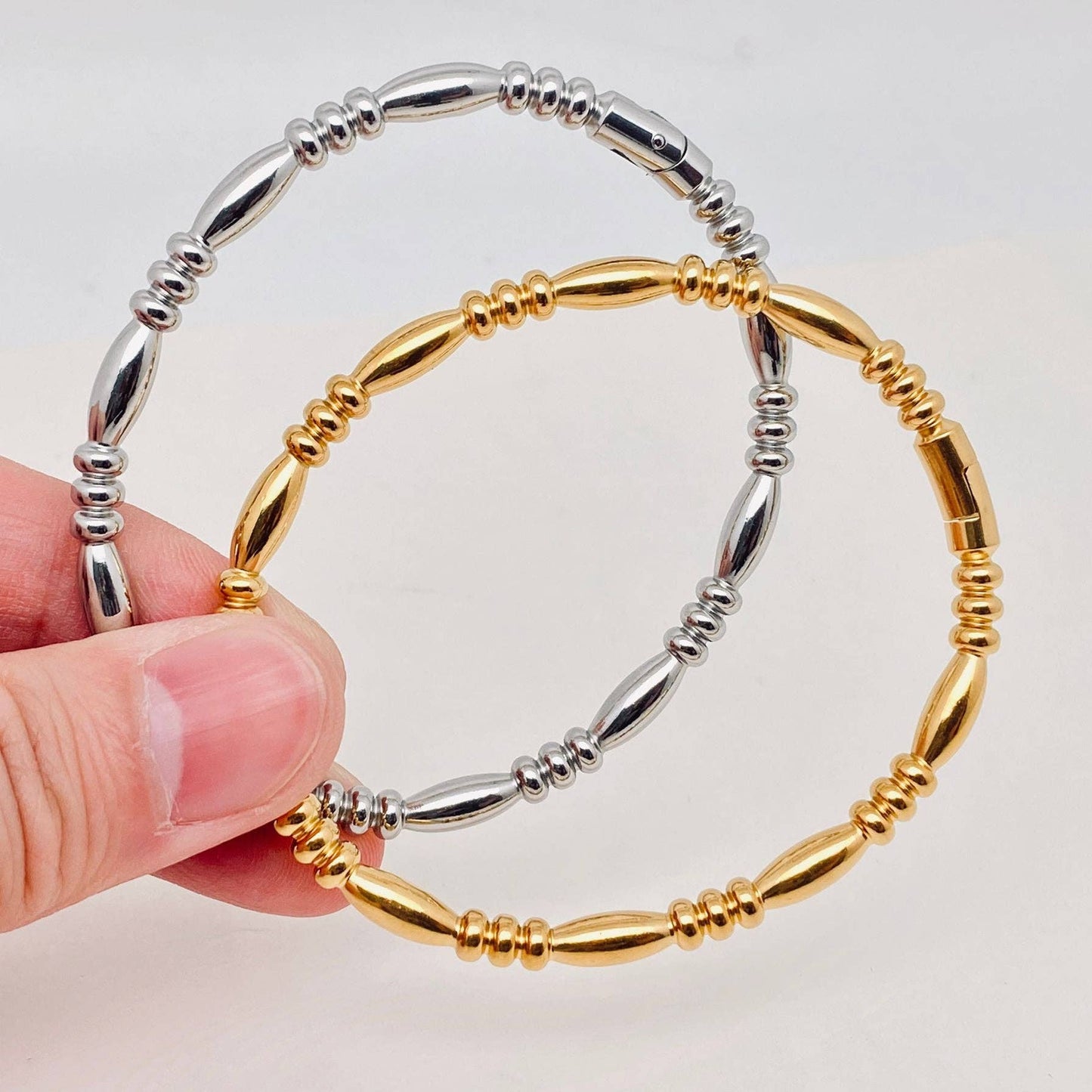 A hand displays two Mio Queena Multi-style 18K Gold-plated Stainless Steel Bangles—one in sleek silver stainless steel and the other in radiant 18K gold plating—with alternating oval and round beads, making them perfect jewelry gifts, set against a plain white background.