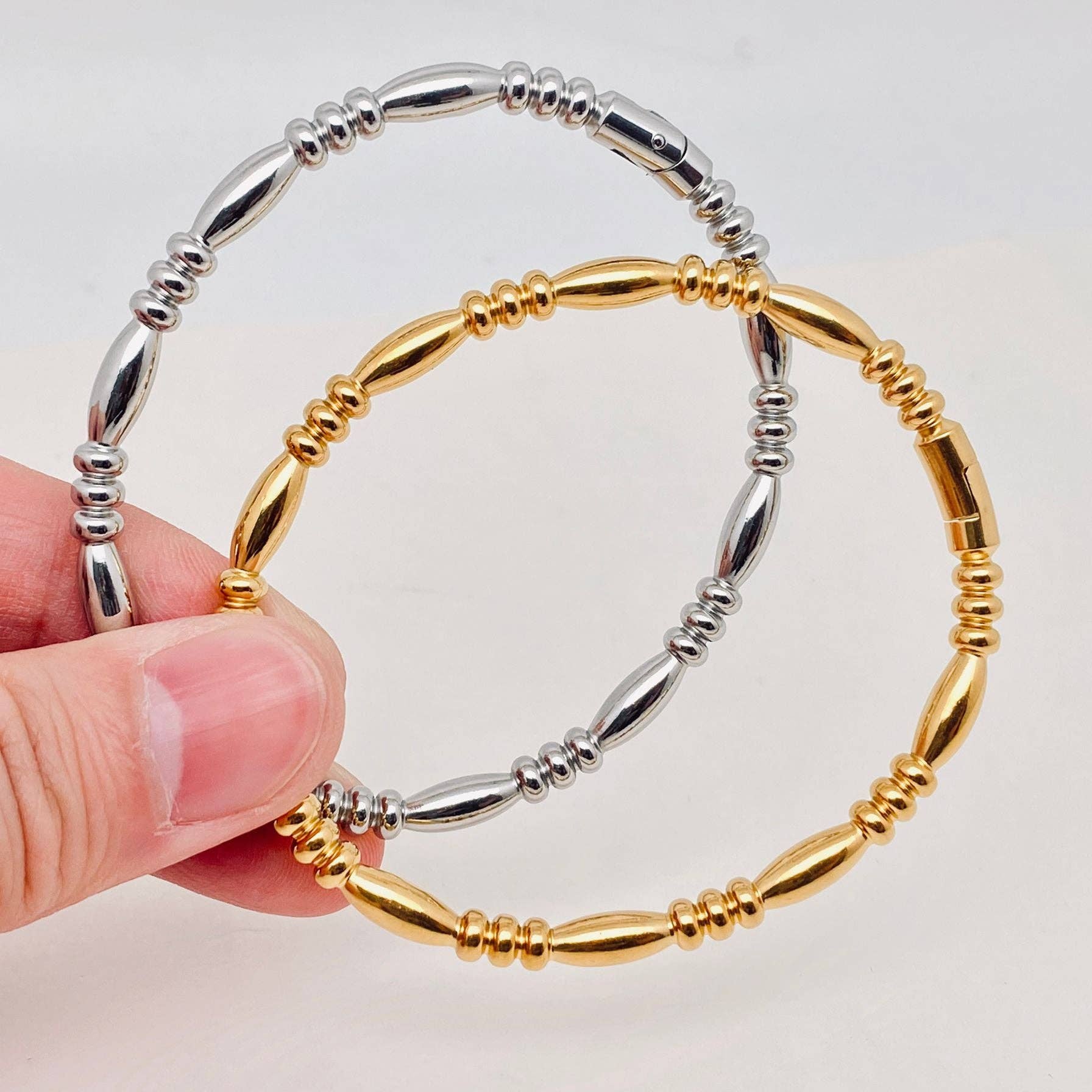 A hand displays two Mio Queena Multi-style 18K Gold-plated Stainless Steel Bangles—one in sleek silver stainless steel and the other in radiant 18K gold plating—with alternating oval and round beads, making them perfect jewelry gifts, set against a plain white background.