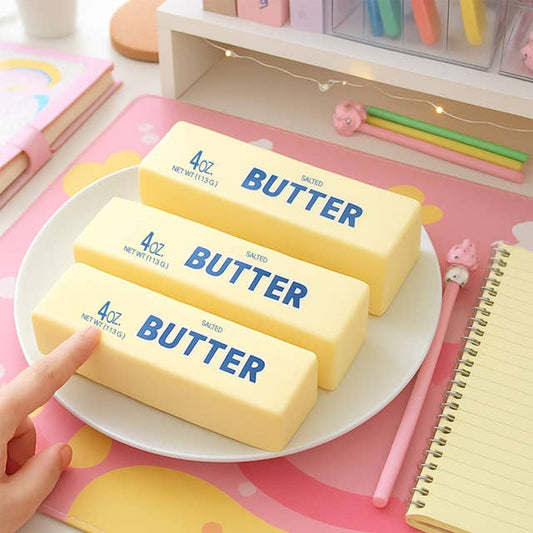 Three yellow butter stick squishies with blue BUTTER text on white plate in colorful desk workspace