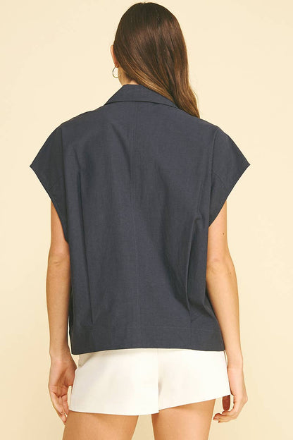 A woman with long brown hair is shown from the back, wearing the PINCH Solid Cap Sleeve Blouse Top in navy and white shorts, standing against a beige background.