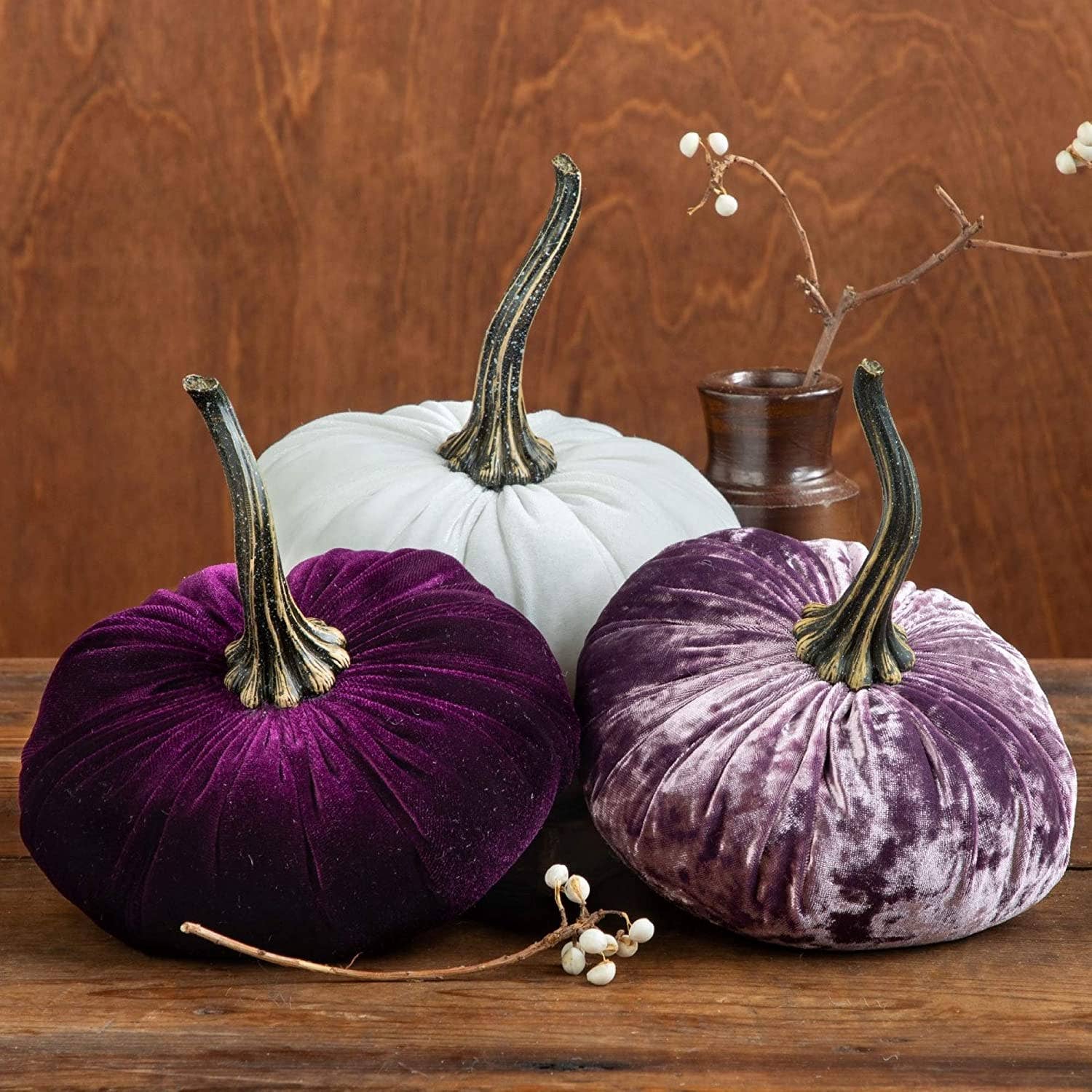 Three large velvet pumpkins from Your Heart’s Content—white, dark purple, and lavender—each featuring a long stem, are arranged on a wooden surface alongside a small brown vase and some sprigs of white berries in the background, creating an elegant Thanksgiving centerpiece or charming holiday mantel décor.