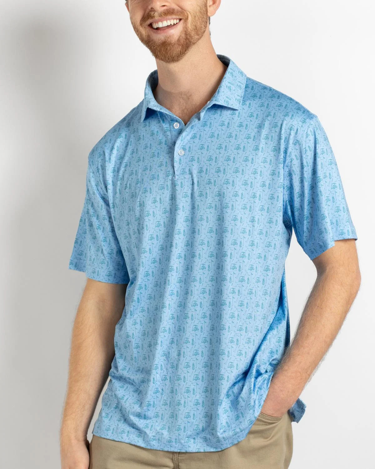 A smiling man with a short beard wears The Royal Standard Men’s Tee Off Performance Polo in a light blue pattern and tan pants. His left hand is in his pocket as he stands against a plain white background.