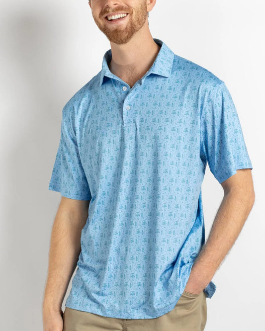 A smiling man with a short beard wears The Royal Standard Men’s Tee Off Performance Polo in a light blue pattern and tan pants. His left hand is in his pocket as he stands against a plain white background.