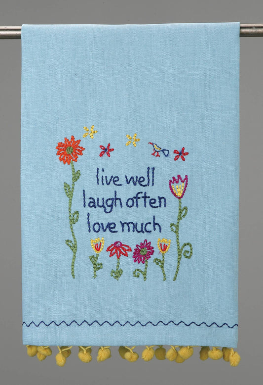Peking Handicraft Live Well Pom Pom Kitchen Towel in light blue with embroidered text and colorful flowers, yellow pom pom trim