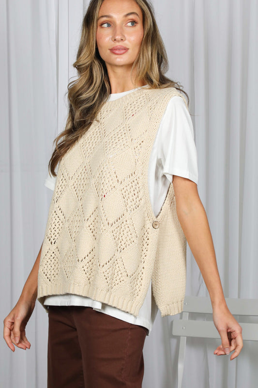 A woman stands in front of white curtains, wearing the Vine & Love A Cozy Textured Sweater Vest in a cream hue over a white t-shirt and brown pants, with a white chair partially visible behind her.