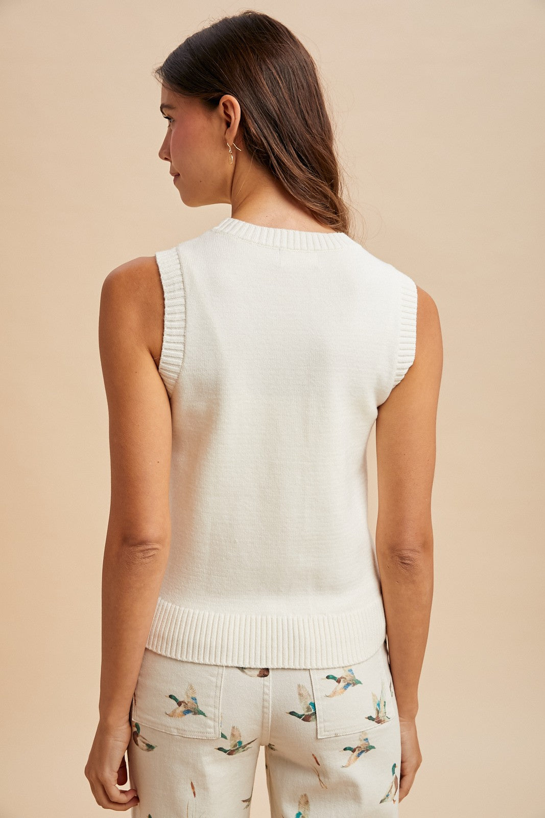 A woman with long brown hair faces away, wearing the Anniewear AnnieWear Mallard Duck Sweater Top— a sleeveless white sweater— paired with cream pants featuring colorful bird prints. The background is plain beige.