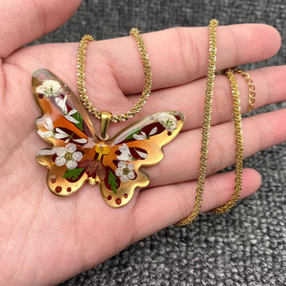 A hand holds the Mio Queena - Dried Flower Brass Butterfly 18K Golden SS Necklace, which features a butterfly-shaped resin pendant encasing delicate dried flowers and leaves. The background is a textured gray fabric.