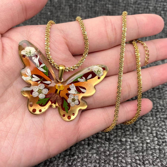 A hand holds the Mio Queena - Dried Flower Brass Butterfly 18K Golden SS Necklace, which features a butterfly-shaped resin pendant encasing delicate dried flowers and leaves. The background is a textured gray fabric.