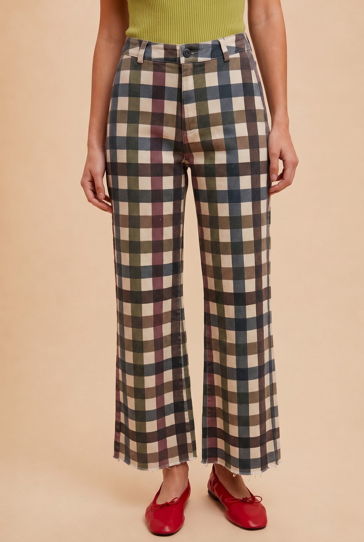 A person wearing the AnnieWear Comfort Stretch Gingham Wide Leg Jean by Anniewear—featuring a high rise, wide-leg silhouette in earthy plaid tones—paired with a green ribbed top and red shoes, stands against a beige background.