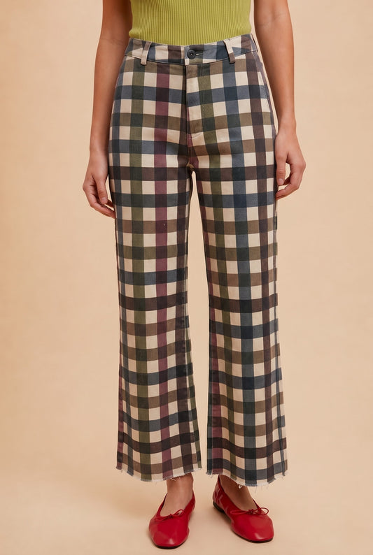A person wearing the AnnieWear Comfort Stretch Gingham Wide Leg Jean by Anniewear—featuring a high rise, wide-leg silhouette in earthy plaid tones—paired with a green ribbed top and red shoes, stands against a beige background.